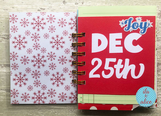 A close-up of a spiral-bound planner or notebook showing two decorated pages side by side. The left page has a white background with a red snowflake pattern, and the right page is bright red with large white text reading 'DEC 25th' and a blue 'Joy' script at the top.