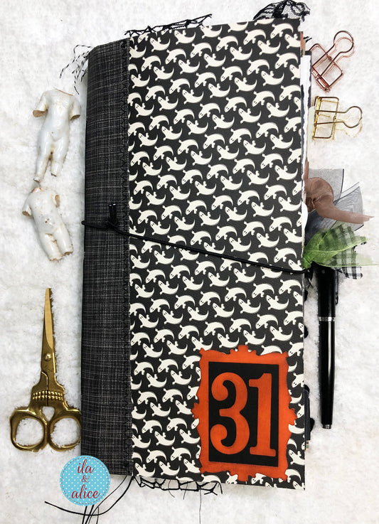 Handmade Halloween junk journal with black and white pattern of ghosts with orange number '31' on a white surface with stationery items.