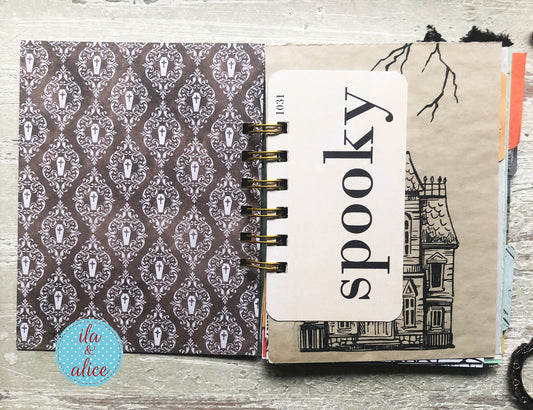 An open spiral-bound notebook or journal on a light wood surface. The left page has a dark brown and gray damask pattern with small vampire illustrations.
