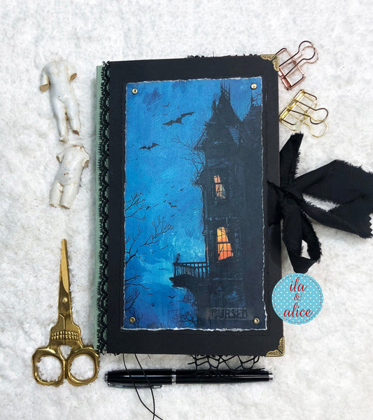 Handmade Eerie Halloween Gothic junk journal with a haunted house illustration on a snowy background, surrounded by stationery items.