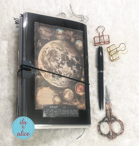 Handmade junk journal with full moon and hare, with celestial design, pen, scissors, and clips on a textured surface.