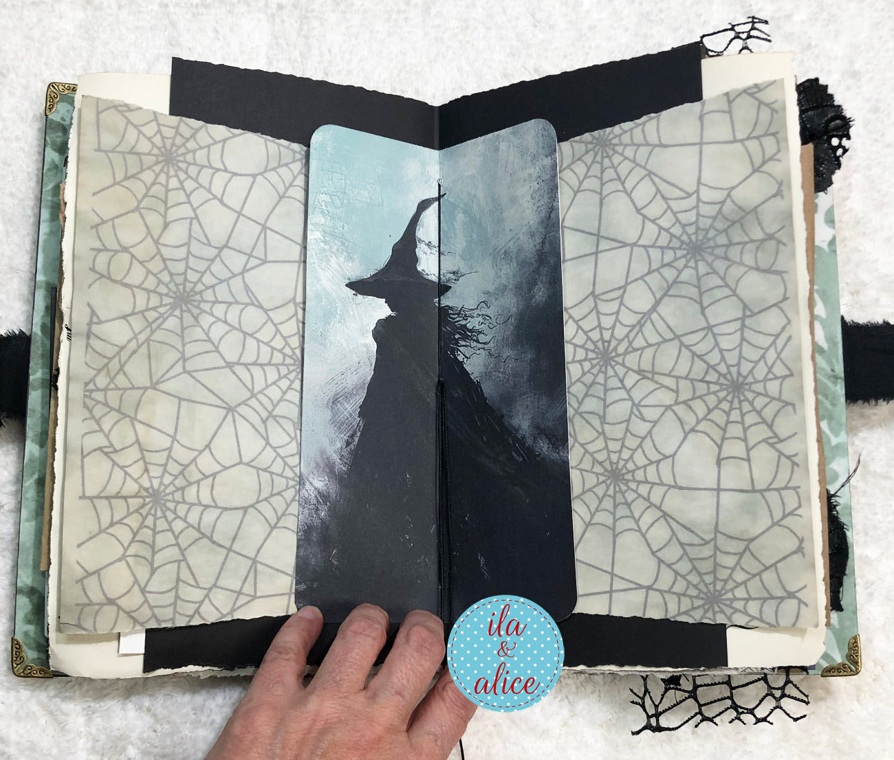 Handmade Eerie Halloween Gothic junk journal with spider web patterns and a silhouette of a witch on a snowy background, with 'ila & alice' branding.