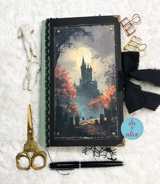 Handmade Eerie Halloween Gothic junk journal with a spooky scene, scissors, pen, and decorative clips on a textured surface.