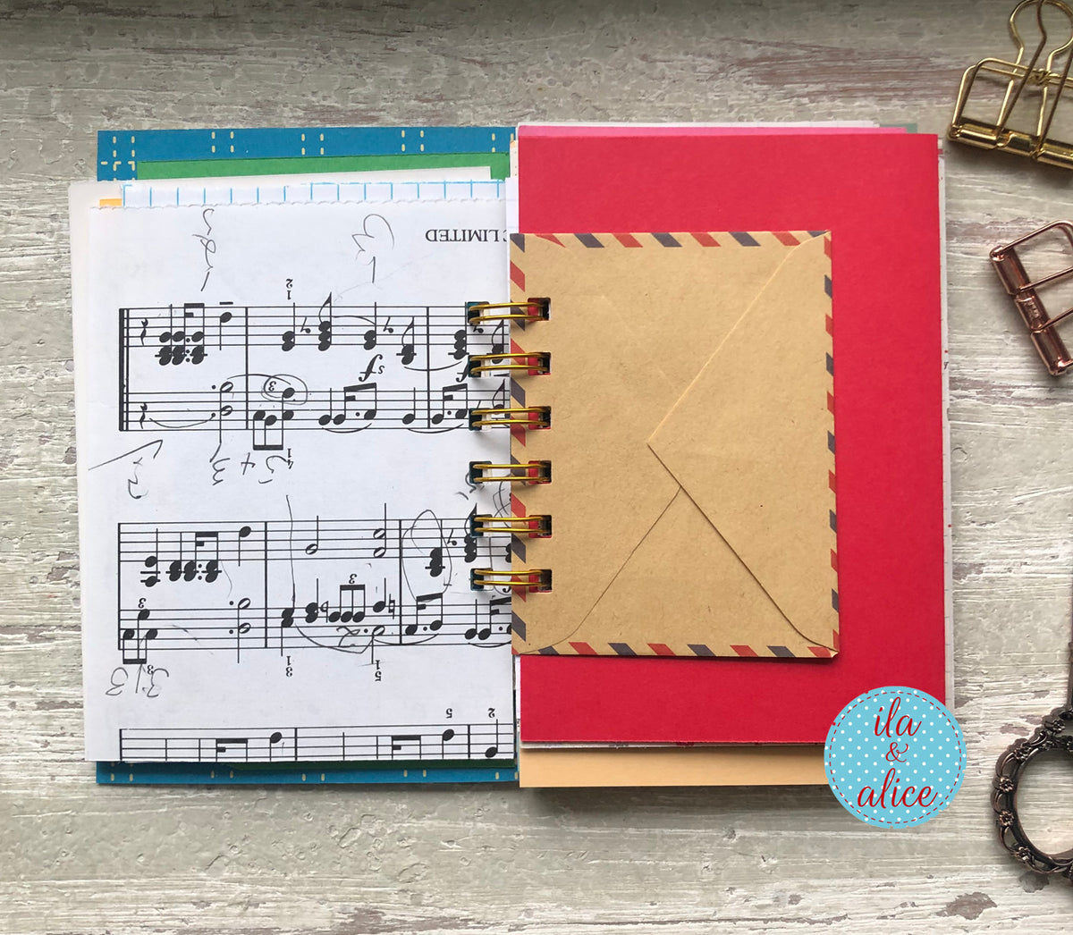 Red notebook with a brown envelope on top, music sheet on the left, on a wooden surface.