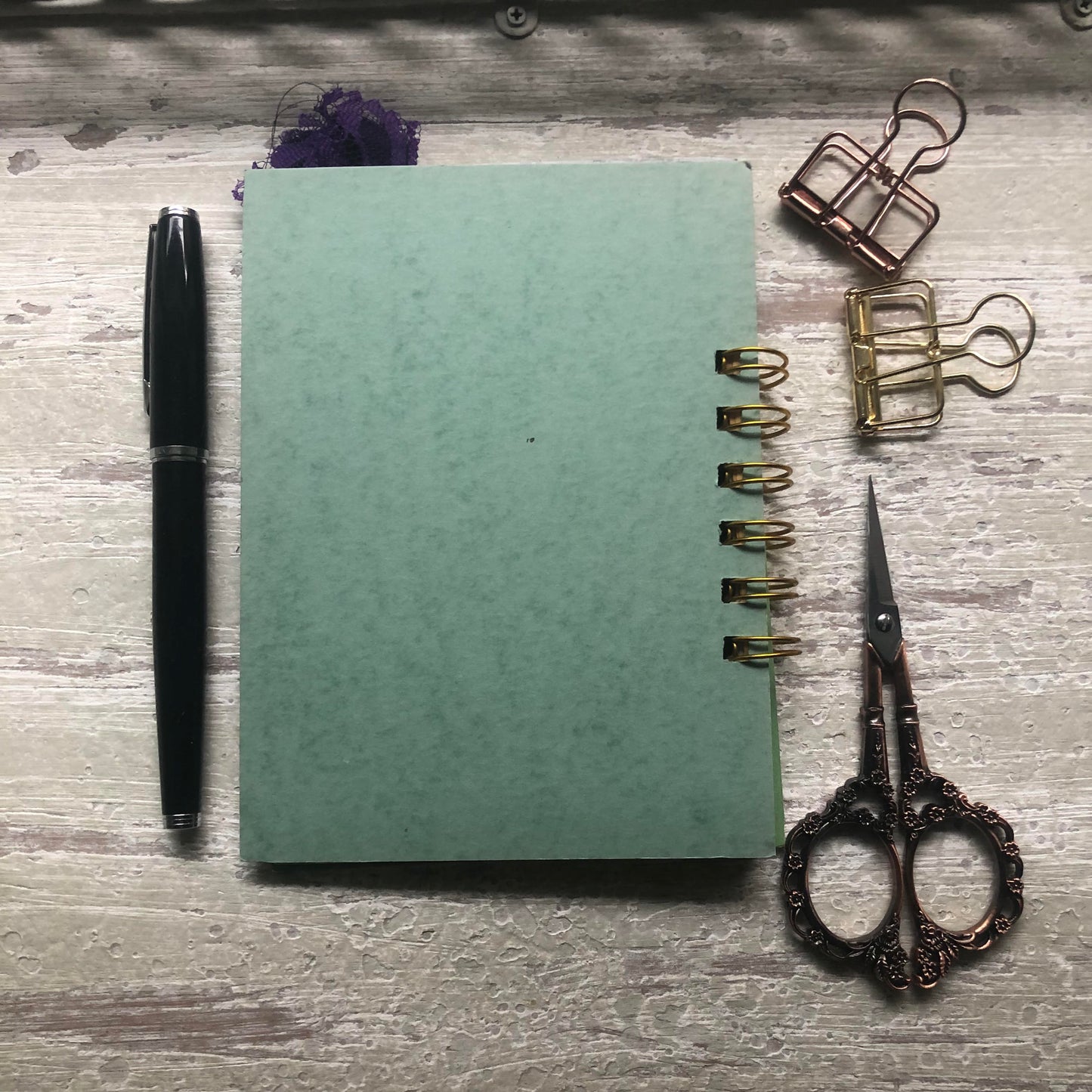 A mint green spiral-bound notebook lies on a distressed gray wooden surface, styled with a black pen on the left, gold and bronze binder clips on the upper right, and ornate scissors on the lower right.