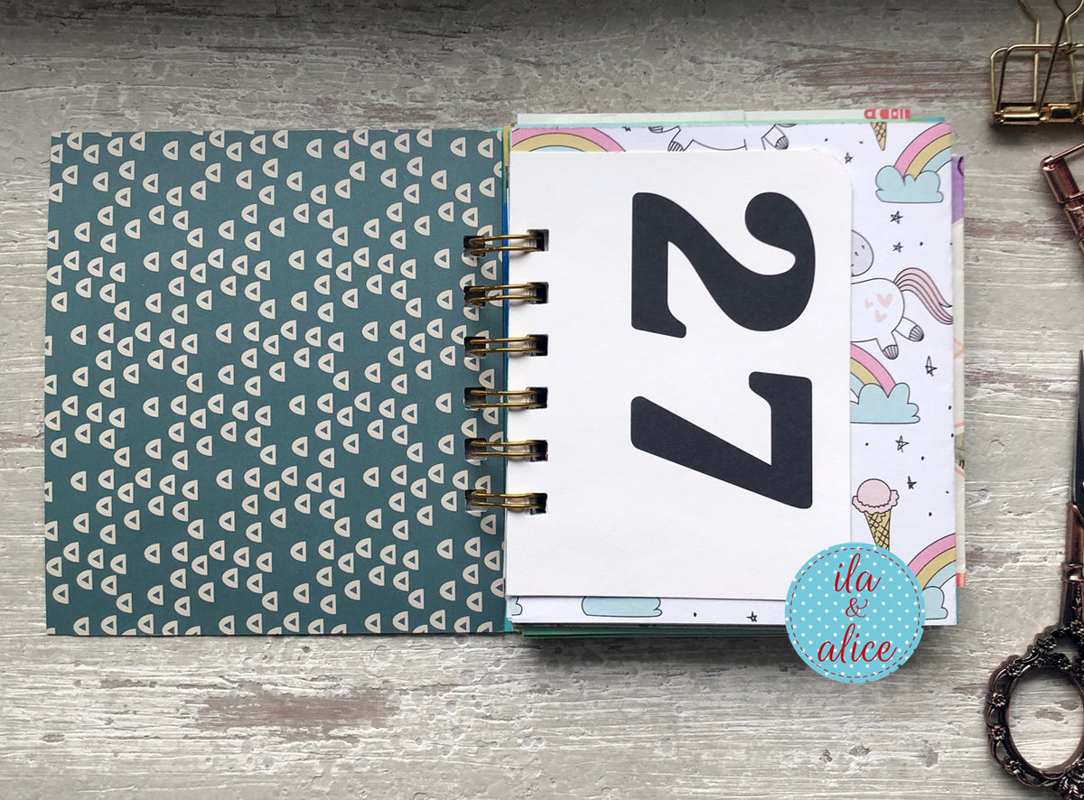 Notebook with a blue geometric pattern on one cover and a white cover with black number '27' and rainbow designs on a wooden surface.