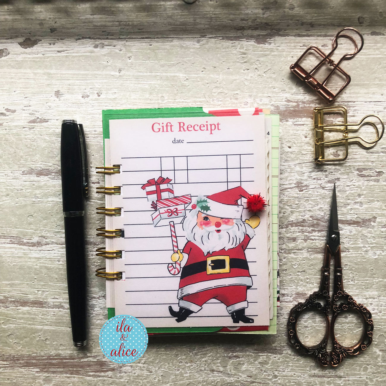 A Christmas-themed gift receipt notepad with a colorful Santa Claus illustration, styled in a flat-lay arrangement on a rustic white-washed wooden background alongside a black pen, gold binder clips, and ornate scissors.
