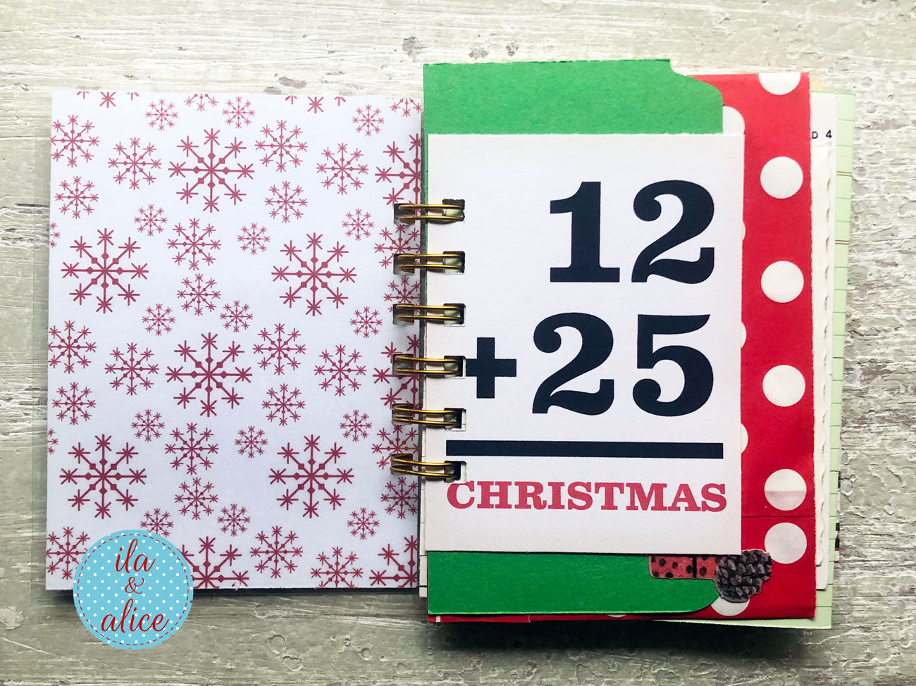 A handmade spiral-bound Christmas advent calendar or planner page set on a light gray wood background. The left page has a white background with red snowflakes, and the right page shows a white insert with black math text reading '12 + 25' and 'CHRISTMAS' in red, accented with green and red-and-white polka dot paper.