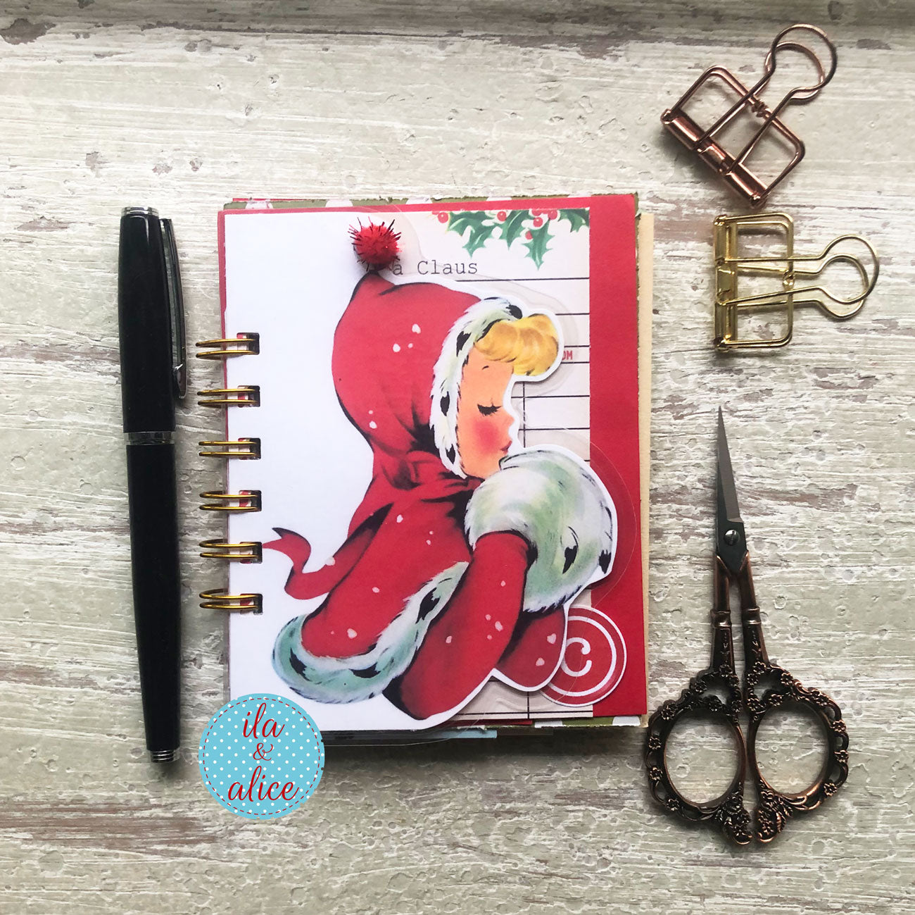 A styled flat-lay of a handmade spiral-bound journal with a vintage Christmas illustration on the cover, surrounded by a black pen, metal paperclips, and ornate scissors on a light wood background.