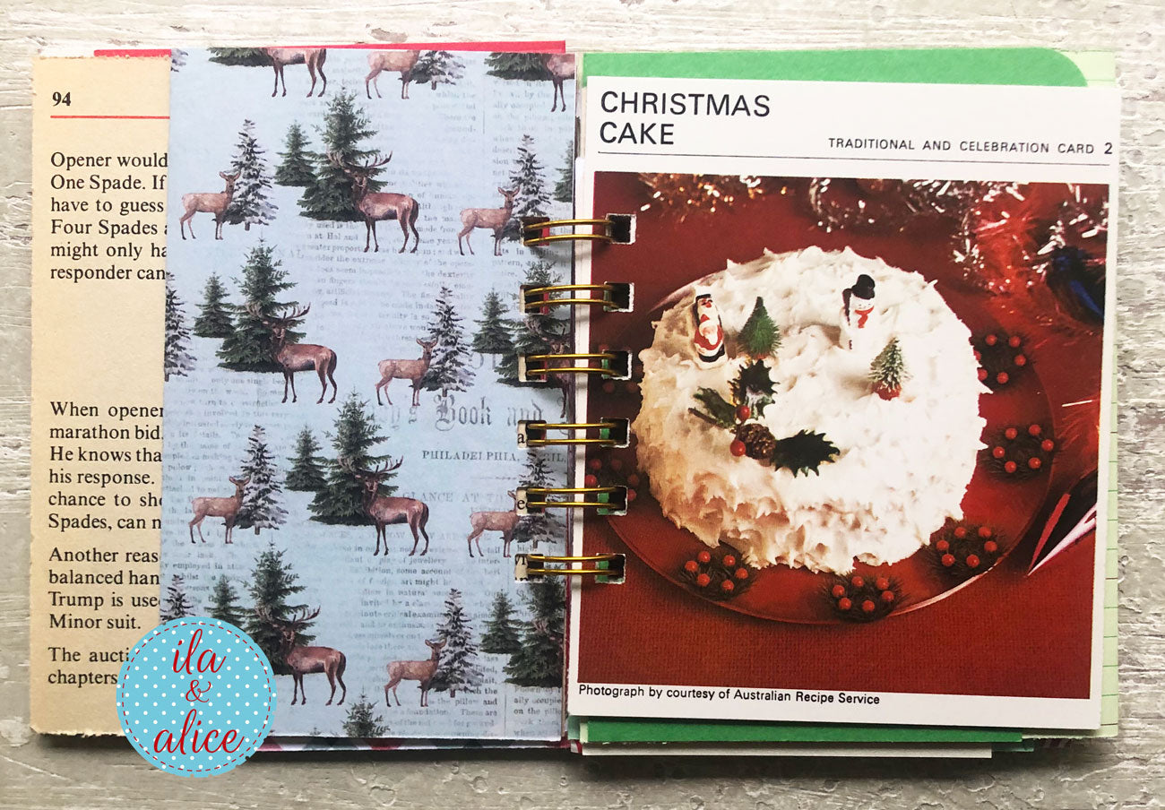 A spiral-bound book or journal spread open on a textured gray surface, showing printed pages with Christmas-themed illustrations, including deer on a tree and a page titled 'Christmas Cake.'.
