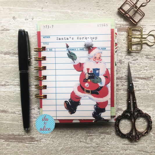 A spiral-bound planner page featuring a vintage illustration of Santa Claus writing in a notebook, styled on a light wood background with a black pen, metal clips, and ornate scissors.