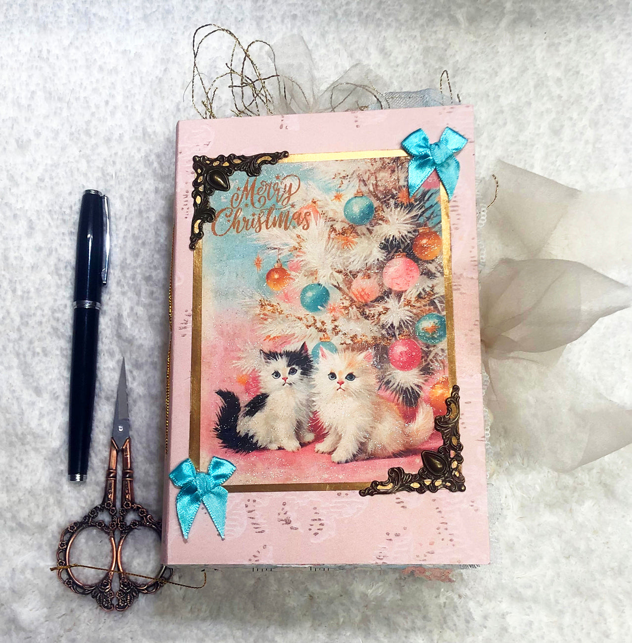 Decorative notebook with Christmas design featuring two cats and ornaments, surrounded by a pen, scissors, and decorative bows on a textured surface.