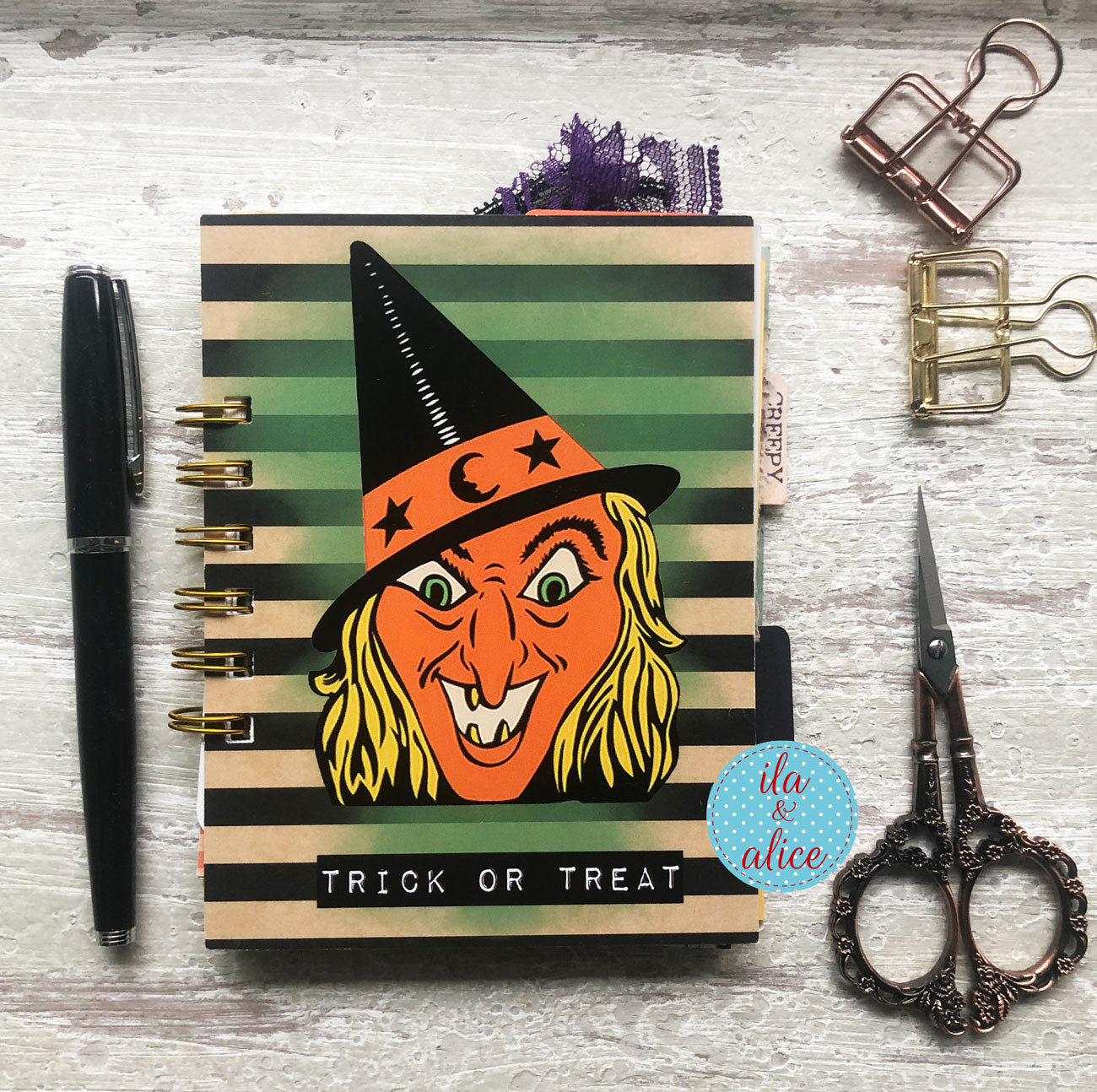 A spiral-bound planner or notebook with a Halloween witch illustration on a striped cover, styled on a whitewashed wood surface with a black pen, metal clips, scissors, and a blue circular sticker.