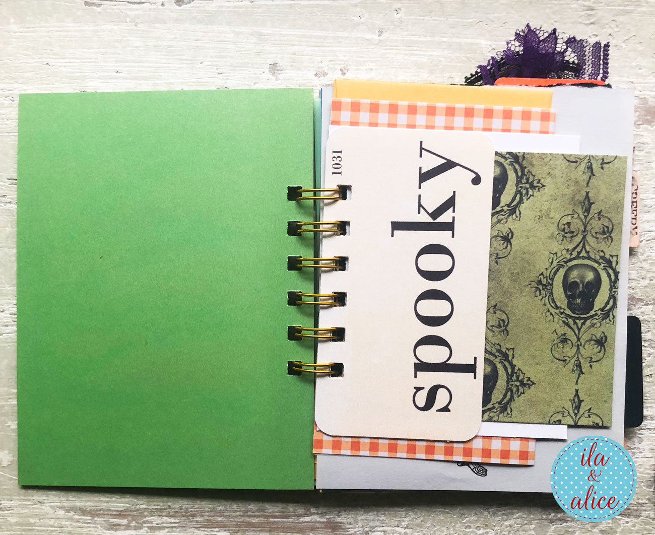 A handmade spiral-bound notebook or planner page with a solid green cover on the left and a decorated insert page on the right featuring the word 'spooky' in black script, orange gingham tape, a vintage skull pattern, and a blue circular 'ila & alice' logo in the bottom right corner.