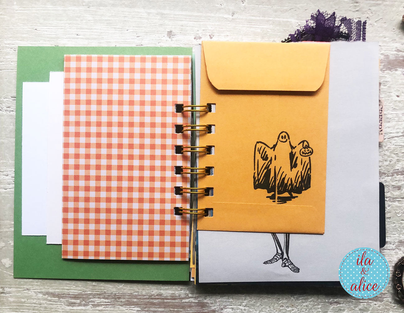 An open spiral-bound notebook or planner on a light wood surface, featuring layered paper inserts including a green card, a white sheet, an orange-and-white gingham sheet, and a yellow envelope with a black ghost illustration. A small blue circular 'ita & alice' logo appears in the bottom right corner.