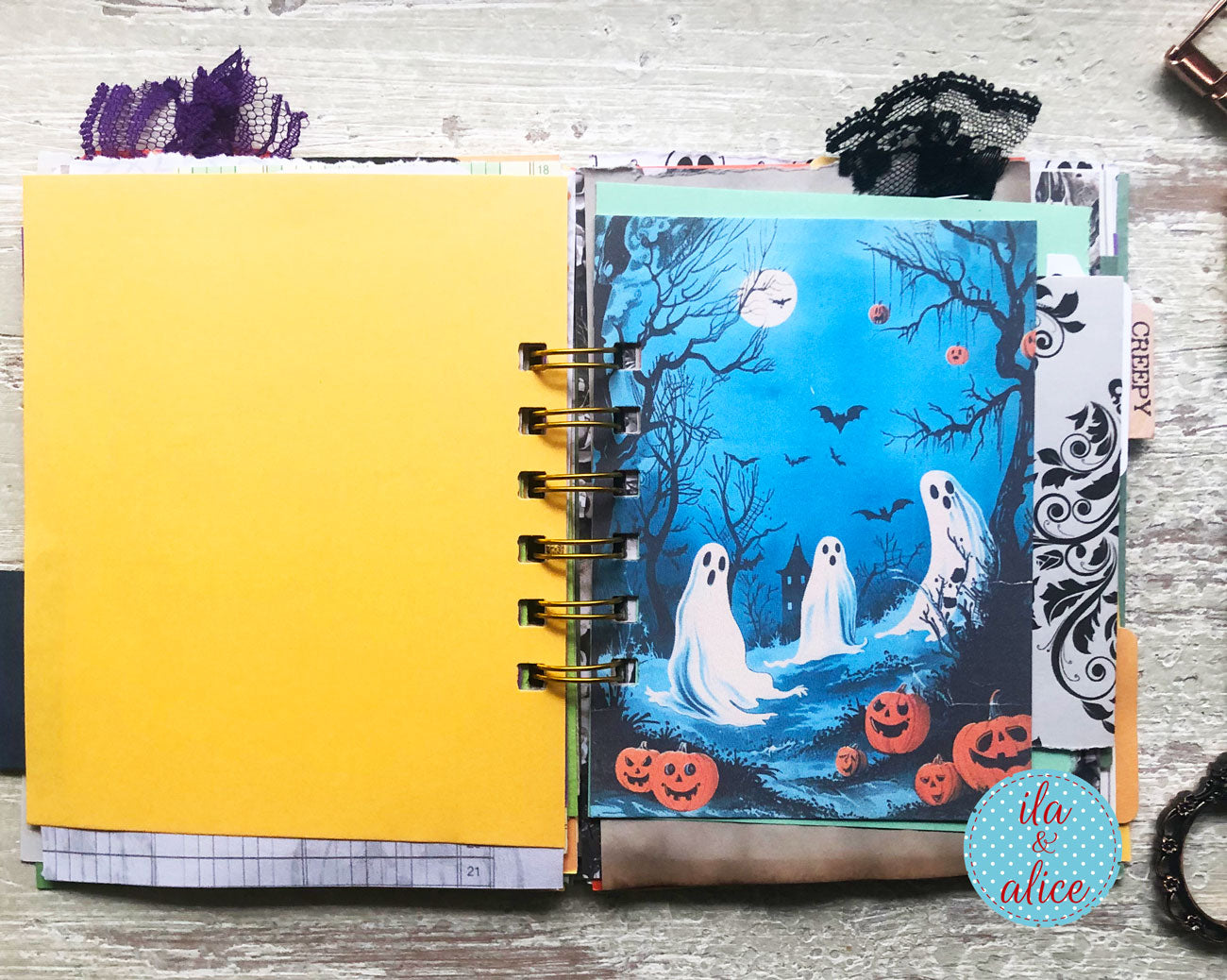 A handmade spiral-bound journal or planner insert featuring a bright yellow left page and a decorated right page with a Halloween illustration of three ghosts in a spooky blue forest with pumpkins, bats, and a full moon. Decorative black lace and patterned tabs are tucked around it on a whitewashed wood surface.