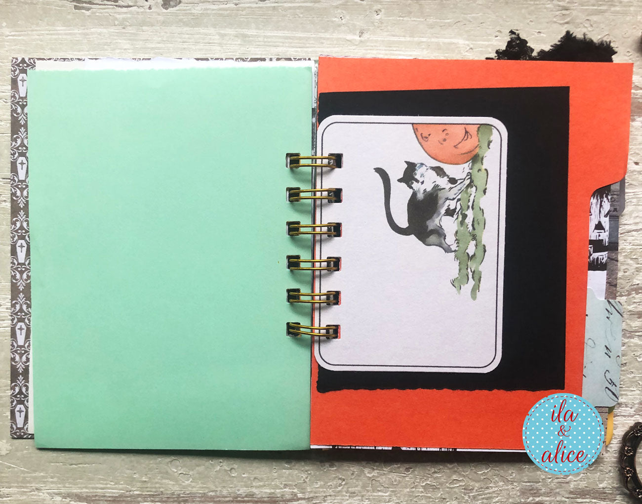 A handmade spiral-bound notebook or journal shown open on a light wood surface. The left page is a solid mint green, while the right page features a layered collage with a black background, a bright orange card, a white insert with a black-and-white cat and orange moon illustration, and decorative paper tabs.