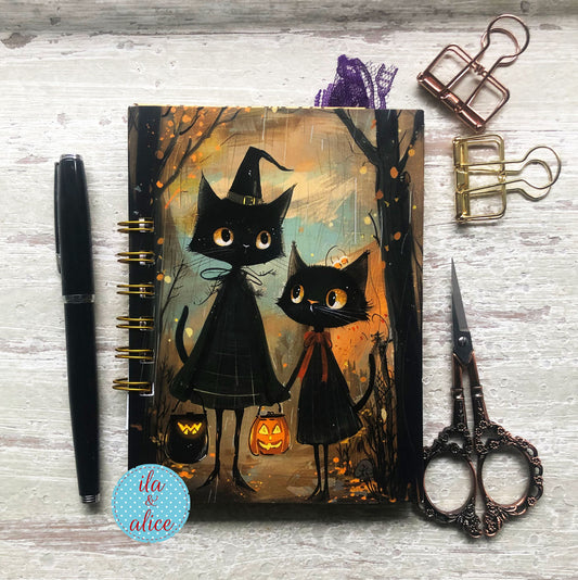 A spiral-bound planner or notebook with a Halloween illustration of two black cats in witch hats, styled on a whitewashed wood surface with a black pen, metal paperclips, ornate scissors, and a blue circular sticker.