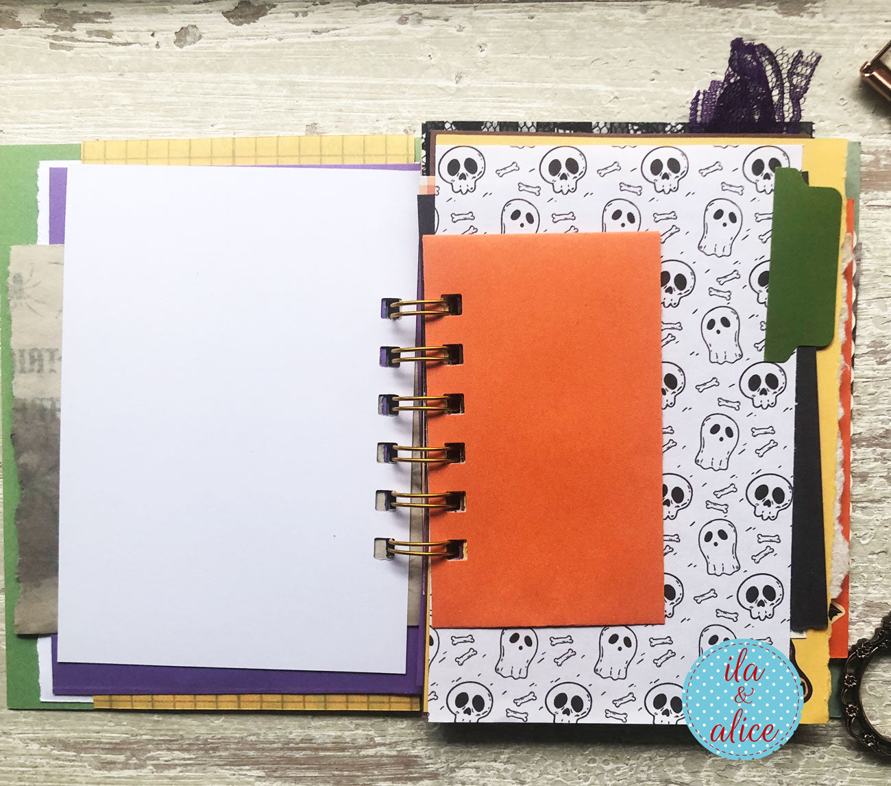 An open handmade spiral notebook with a gold wire binding, featuring a white page on the left and a decorated cover on the right with a pattern of black skulls and bones, a bright orange pocket, and various decorative paper tabs.