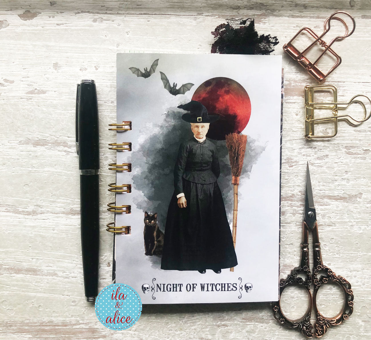 A spiral-bound notebook with a 'Night of Witches' witch illustration, styled on a white wood background with a black pen, metal clips, scissors, and a decorative sticker.