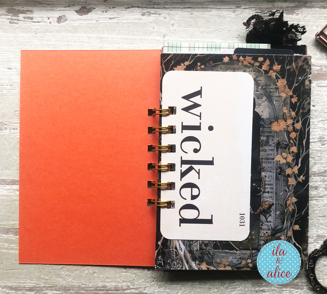 A spiral-bound notebook or journal cover laid flat on a distressed white wood surface. The cover has a large white label reading 'wicked' in black serif text, with a black-and-brown floral/ornamental background and gold spiral binding.