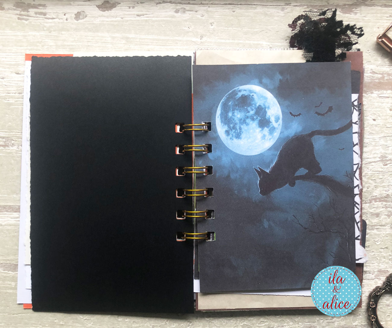 An open spiral-bound notebook or journal on a light wood surface, featuring a black cover on the left and a printed page on the right with a full moon, a cat silhouette, and bats. A small circular 'ila & alice' logo appears in the bottom right corner.