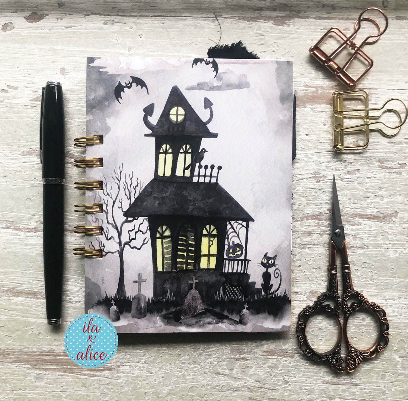 A spiral-bound notebook with a Halloween haunted-house illustration, styled on a whitewashed wood surface with a black pen, brass binder clips, and ornate scissors.