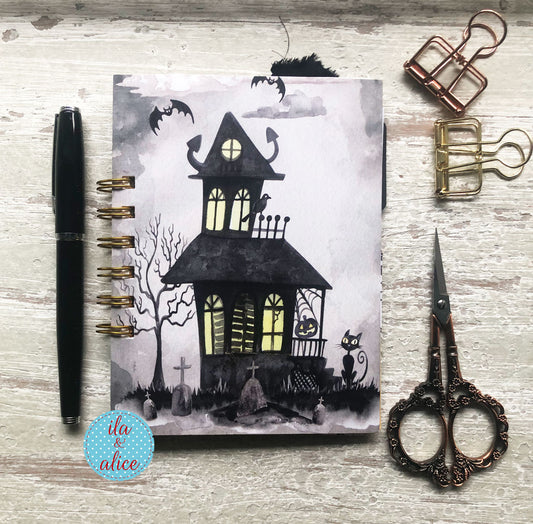 A spiral-bound notebook with a Halloween haunted-house illustration, styled on a whitewashed wood surface with a black pen, brass binder clips, and ornate scissors.