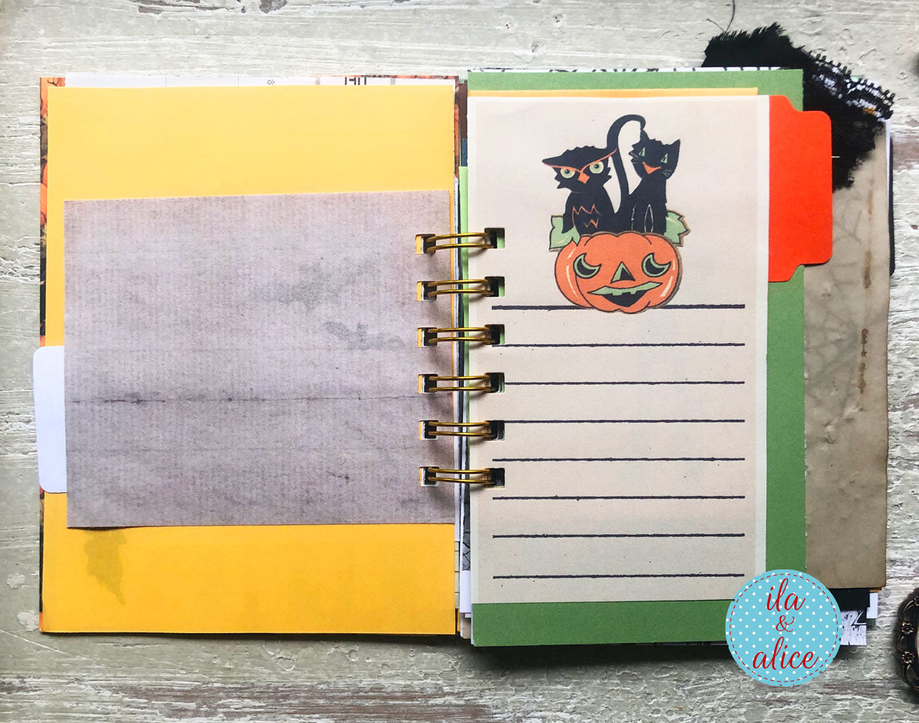 A handmade spiral-bound planner or notebook with a yellow cover, a gray textured insert, and a lined page decorated with a black cat and pumpkin illustration. The notebook is displayed on a rustic gray wooden surface.