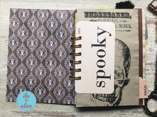 An open spiral-bound notebook or journal laid flat on a rustic wood surface. The left page has a dark brown and gray damask pattern with small tuxedo silhouettes.