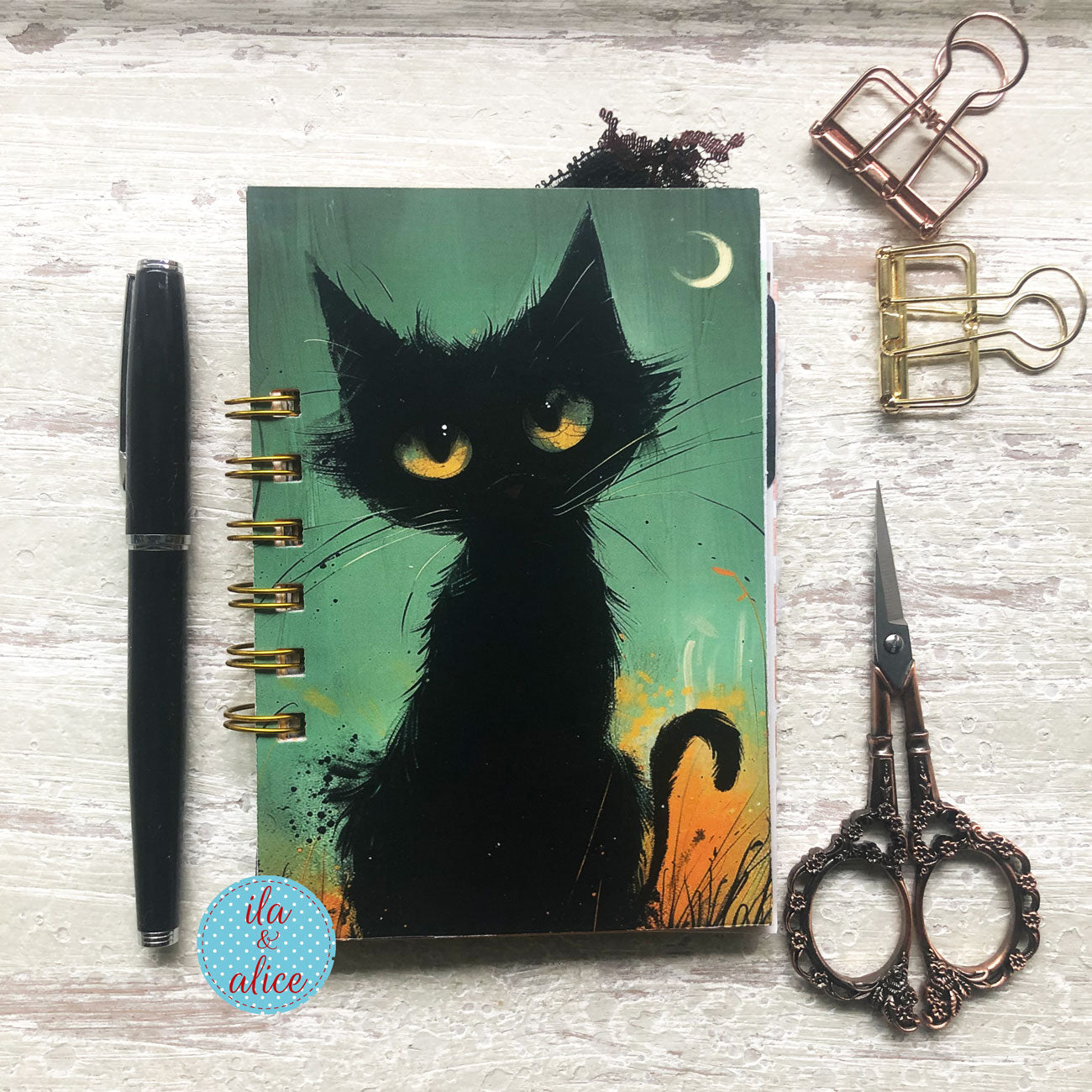 A spiral-bound notebook with a black cat illustration on the cover, styled on a whitewashed wood surface with a black pen, two binder clips, and ornate scissors.