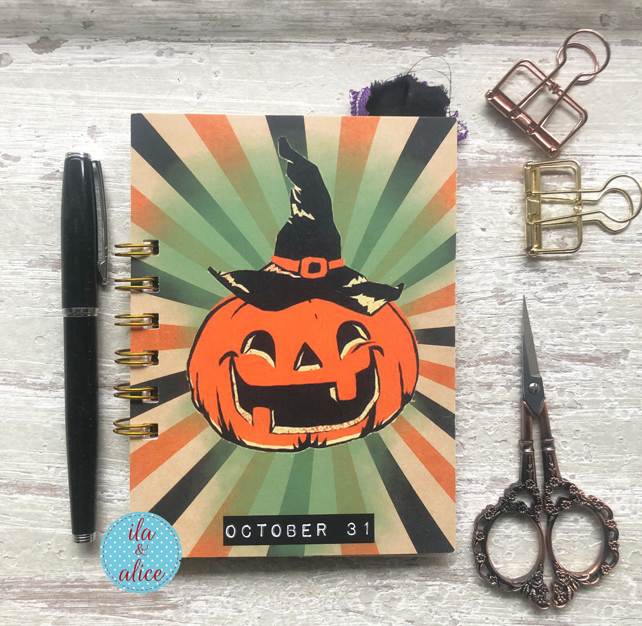 A spiral-bound planner or notebook with a Halloween pumpkin illustration on the cover, placed on a whitewashed wood surface with a black pen, metal clips, and ornate scissors around it.