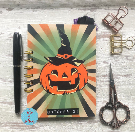 A spiral-bound planner or notebook with a Halloween pumpkin illustration on the cover, placed on a whitewashed wood surface with a black pen, metal clips, and ornate scissors around it.