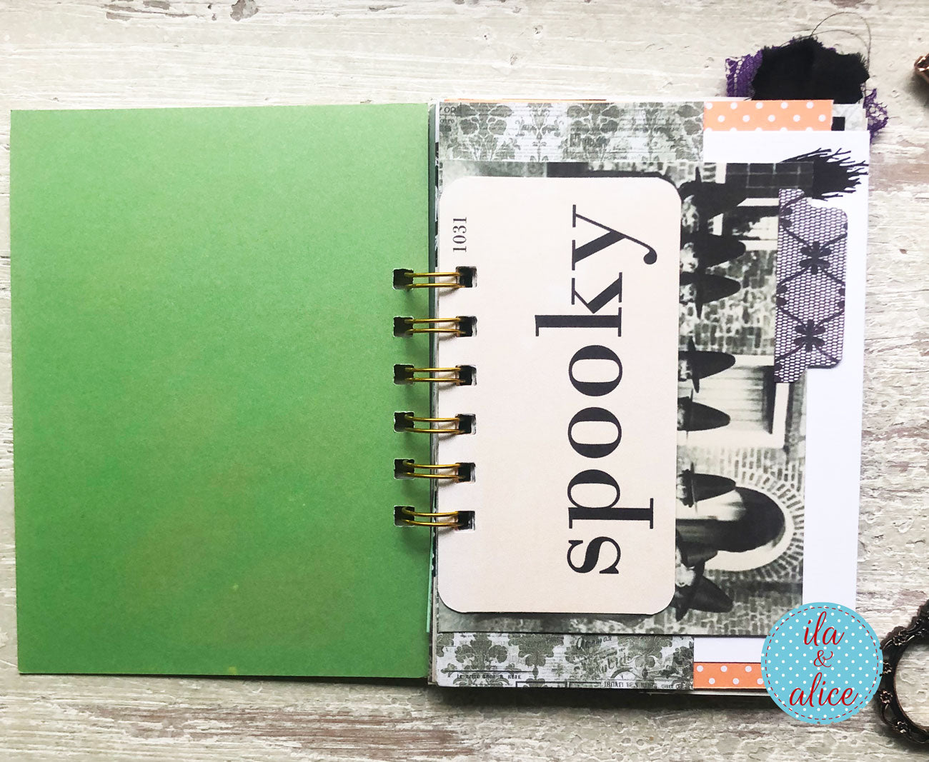A handmade spiral-bound notebook or planner page set on a light wood surface. The left page is solid green, and the right page features a collage with a cream card reading 'spooky' in black script, black-and-white spooky illustrations, patterned paper, and gold spiral binding.