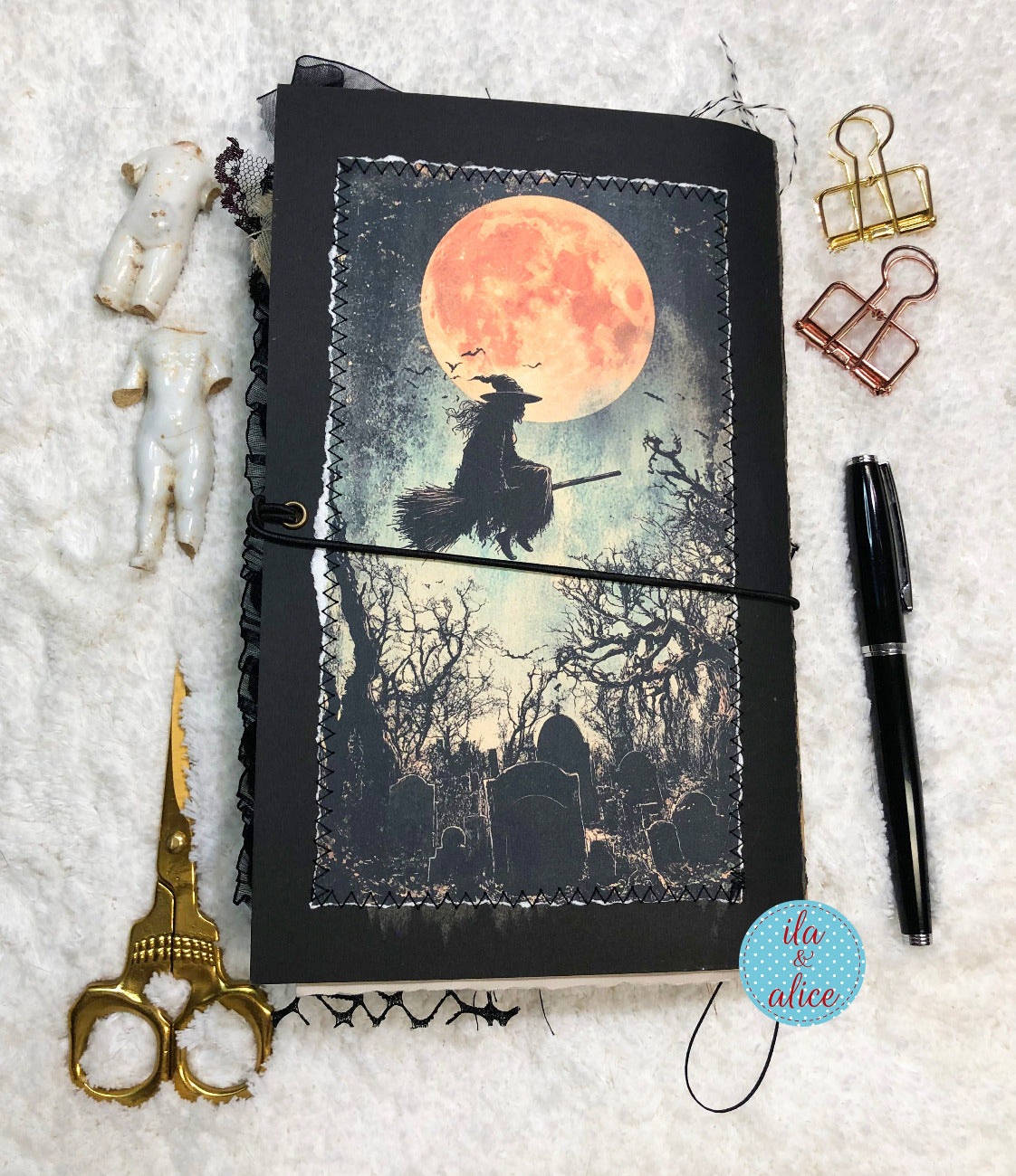 A handmade journal with a gothic witch illustration on the cover, styled in a flat-lay arrangement on a white textured background with gold scissors, white figurines, binder clips, a black pen, and a blue brand sticker reading 'ila alice'.