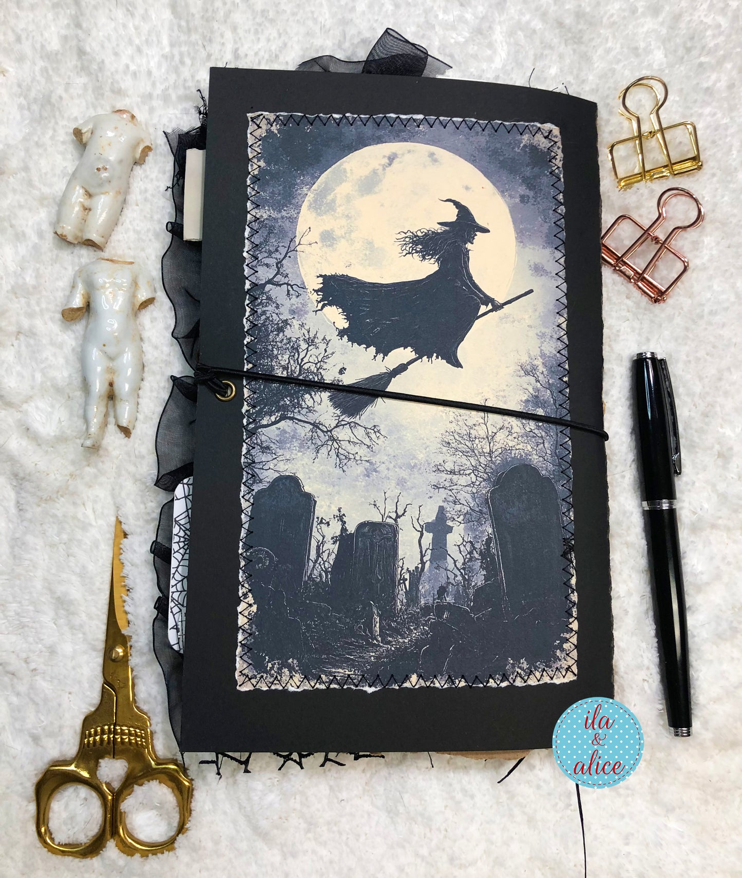 A handmade witch-themed journal with a black cover and a printed illustration of a witch flying on a broomstick in front of a full moon over a graveyard, styled on a white textured background with gold scissors, white figurines, binder clips, a black pen, and a blue 'do & alice' sticker.