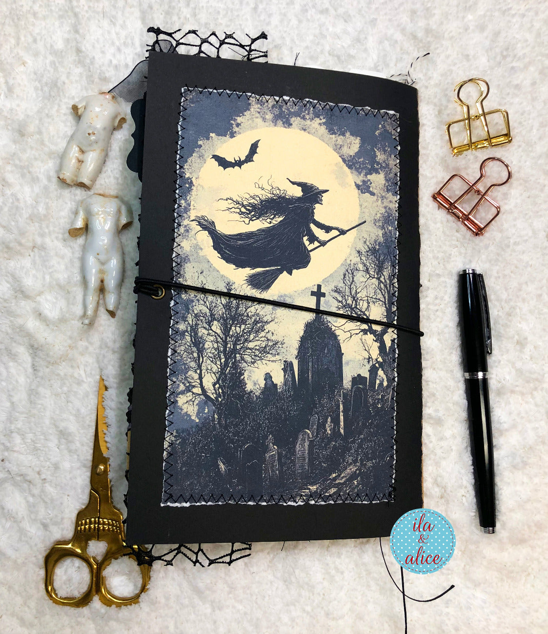 A handmade gothic journal with a black cover and a stitched front cover panel depicting a witch flying over a graveyard under a full moon, styled on a white textured background with scissors, binder clips, figurines, and a pen.