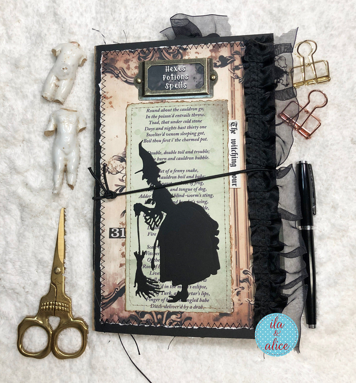 A styled flat-lay of a handmade journal with a witch-themed page, black tulle, and a black ribbon, surrounded by gold scissors, paper clips, a pen, and small white figurines on a white textured fabric background.