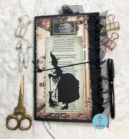 A styled flat-lay of a handmade journal with a witch-themed page, black tulle, and a black ribbon, surrounded by gold scissors, paper clips, a pen, and small white figurines on a white textured fabric background.