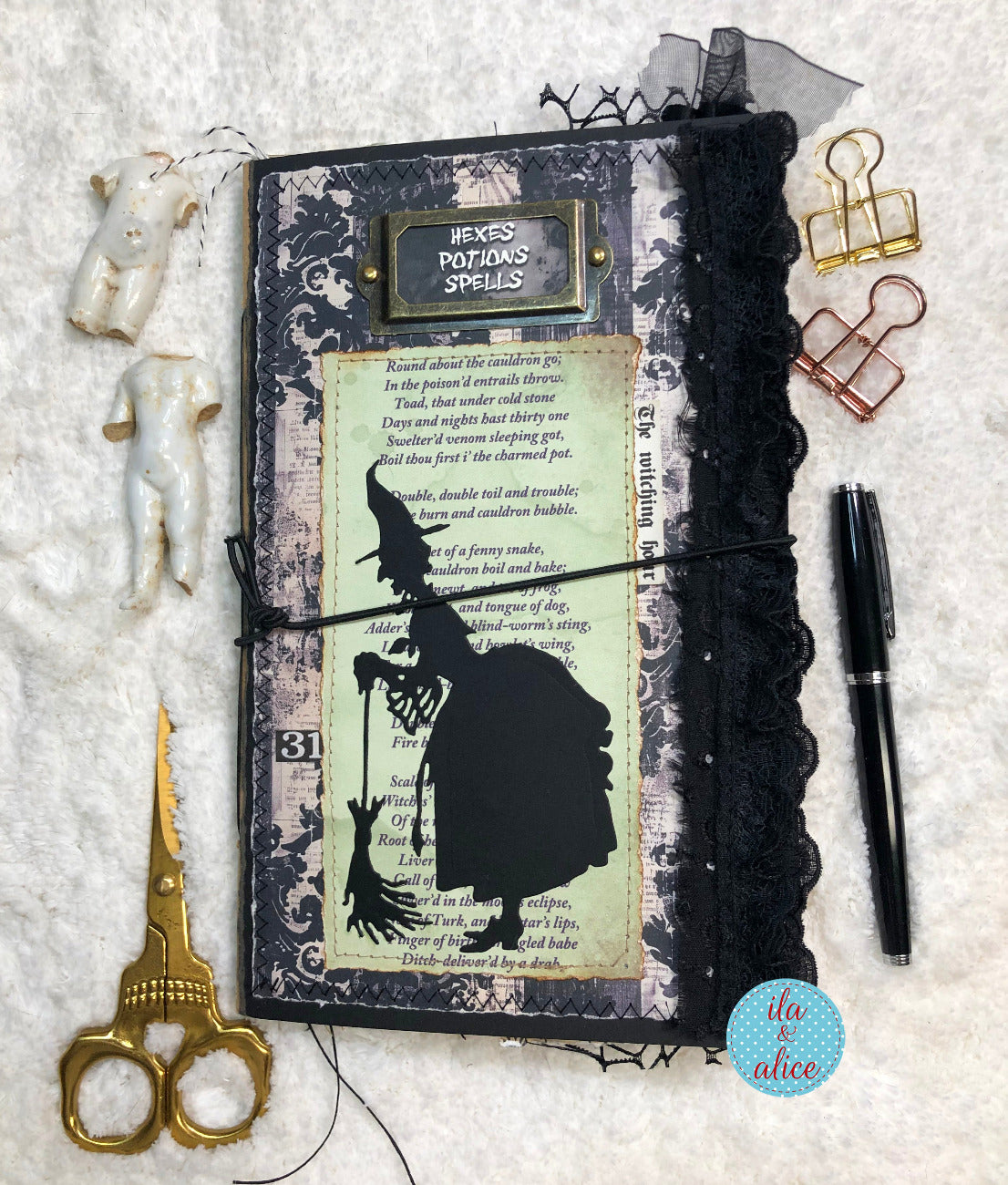 A gothic-themed handmade journal featuring a black silhouette of a witch, vintage spell text, lace trim, and a black elastic band, styled on a white textured background with gold scissors, binder clips, a pen, and small decorative charms.