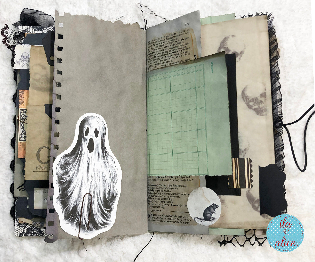 An open handmade scrapbook journal or art book laid flat on a white textured surface. The pages feature vintage-style collage elements including aged paper, black lace trim, handwritten notes, a ghost illustration, a cat sticker, and a pale green grid sheet.