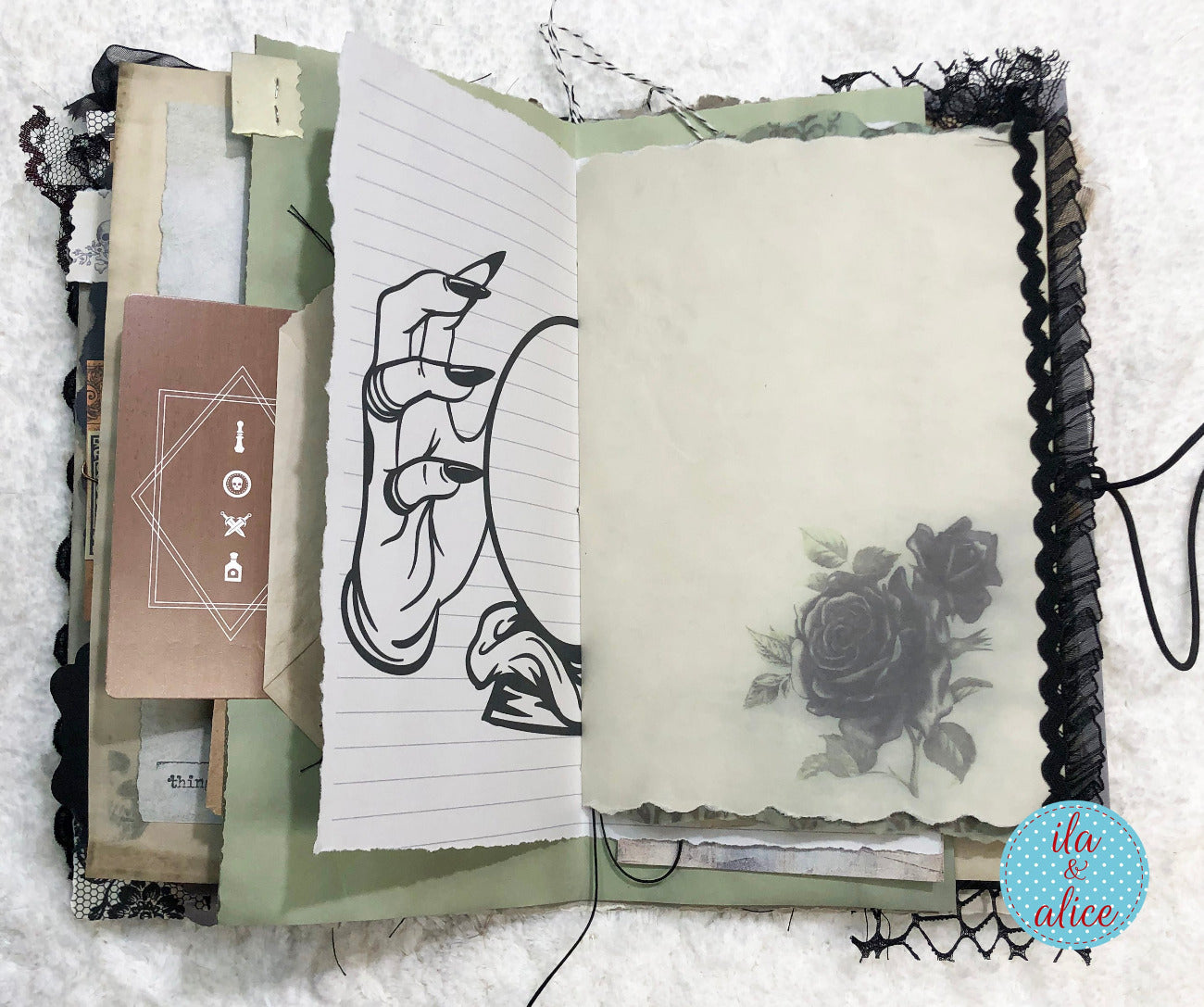 An open handmade paper journal with a distressed, gothic aesthetic. The pages feature a lined sheet with a black ink illustration of a hand holding a mirror, a blank cream page with a vintage-style black rose print, and various layered paper elements, ribbons, and tags.
