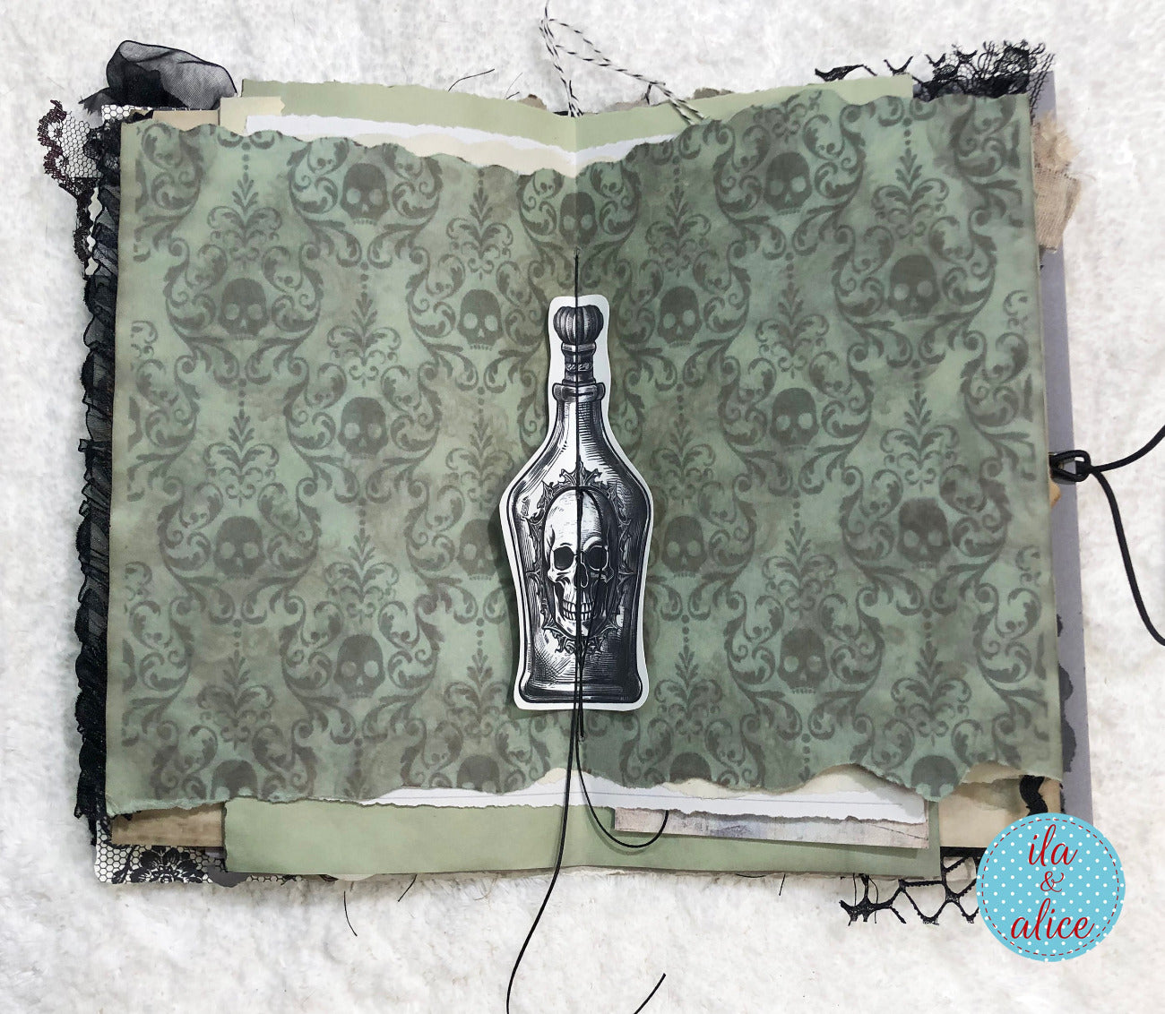 A handmade gothic scrapbook album or journal featuring a central pull-out tab with a skull-and-bottle illustration, set against a patterned sage green and gray damask paper background with black lace trim.