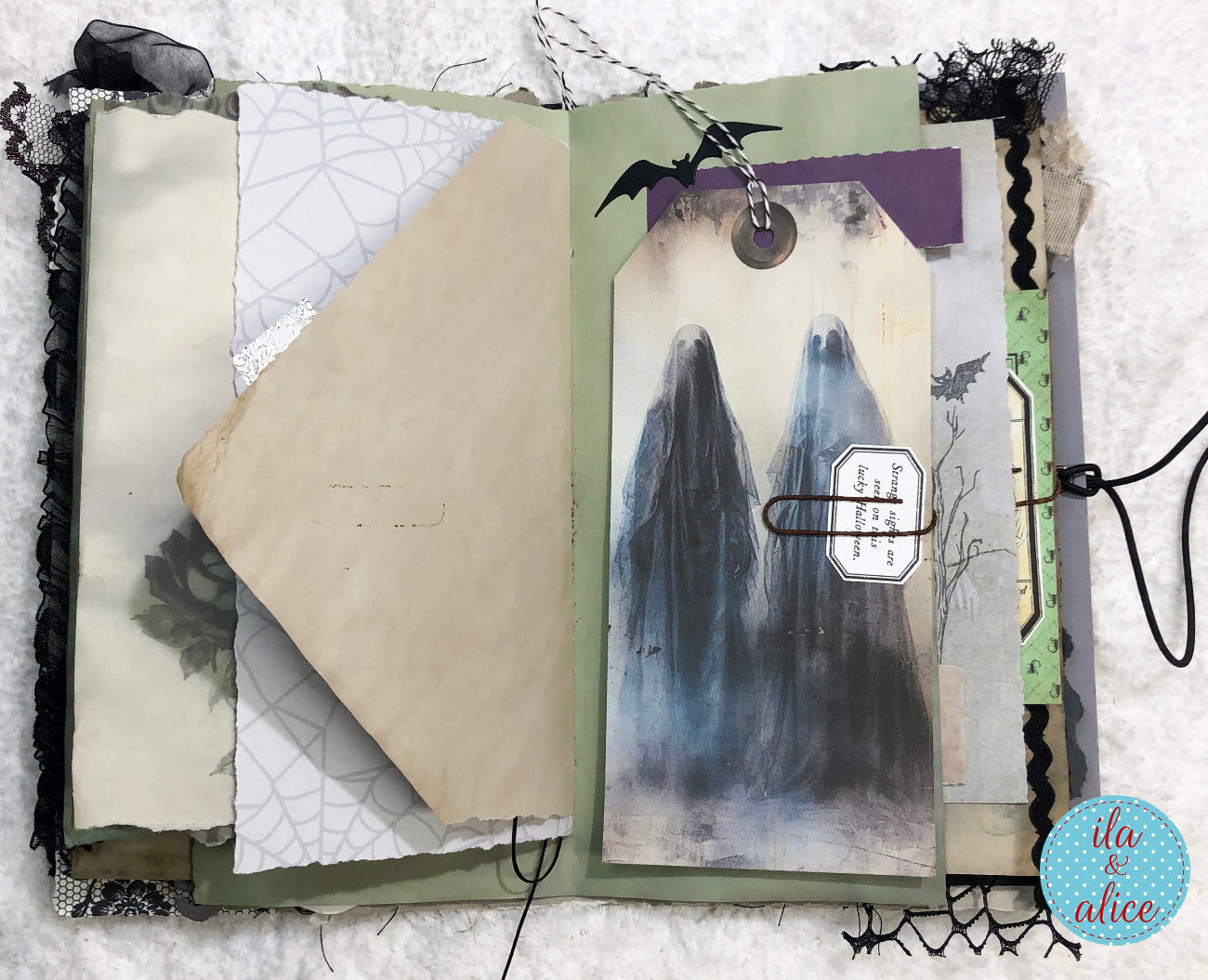 An open handmade scrapbook journal with a spooky ghost-themed page. The page features a watercolor illustration of two ghosts, black lace trim, a vintage-style paper tag, a metal paperclip, and a small bat cutout, all arranged on a white textured fabric background.