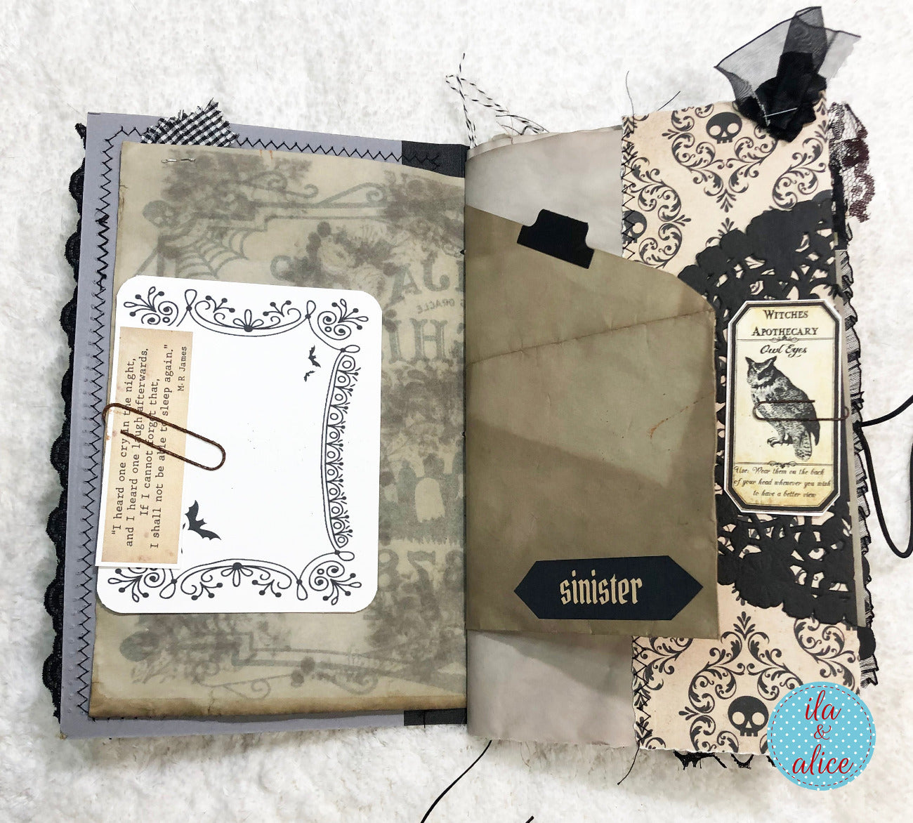 A handmade gothic-themed scrapbook album or journal spread open on a white textured fabric background. The pages feature vintage-style illustrations, black lace overlays, skull motifs, and gothic typography, including the word 'sinister' and a 'Witch's Amulet Card'.