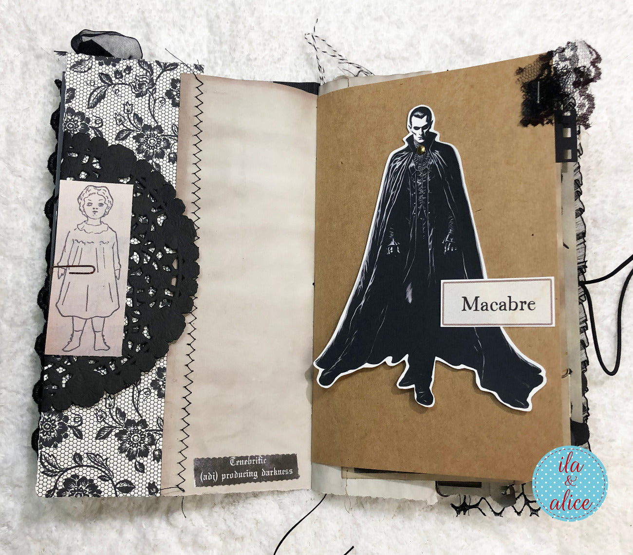 An open handmade scrapbook journal or art book displayed on a white textured fabric background. The left page features black-and-white floral lace, a vintage-style portrait card, and a black lace applique.