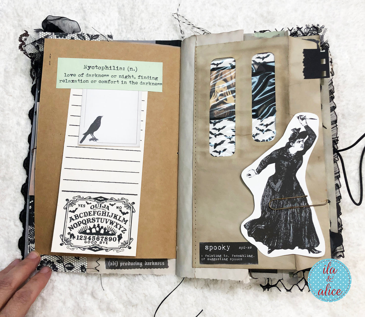 An open handmade paper journal with gothic-themed pages, featuring a brown kraft paper cover, black lace trim, a bird silhouette, an allover Ouija board cutout, and a black-and-white illustration of a dancing woman. A hand is holding the left page, and the background is a white textured fabric.