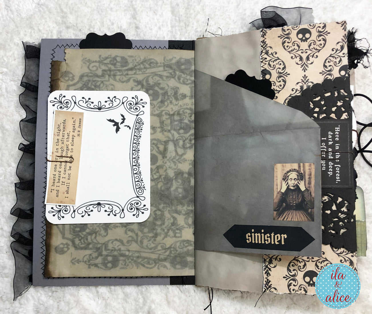 A handmade gothic scrapbook album or journal shown open on a white textured fabric background. The pages feature dark gray, black, and beige tones with skull and bat motifs, layered paper textures, a small vintage-style portrait, and decorative black lace-like trim.