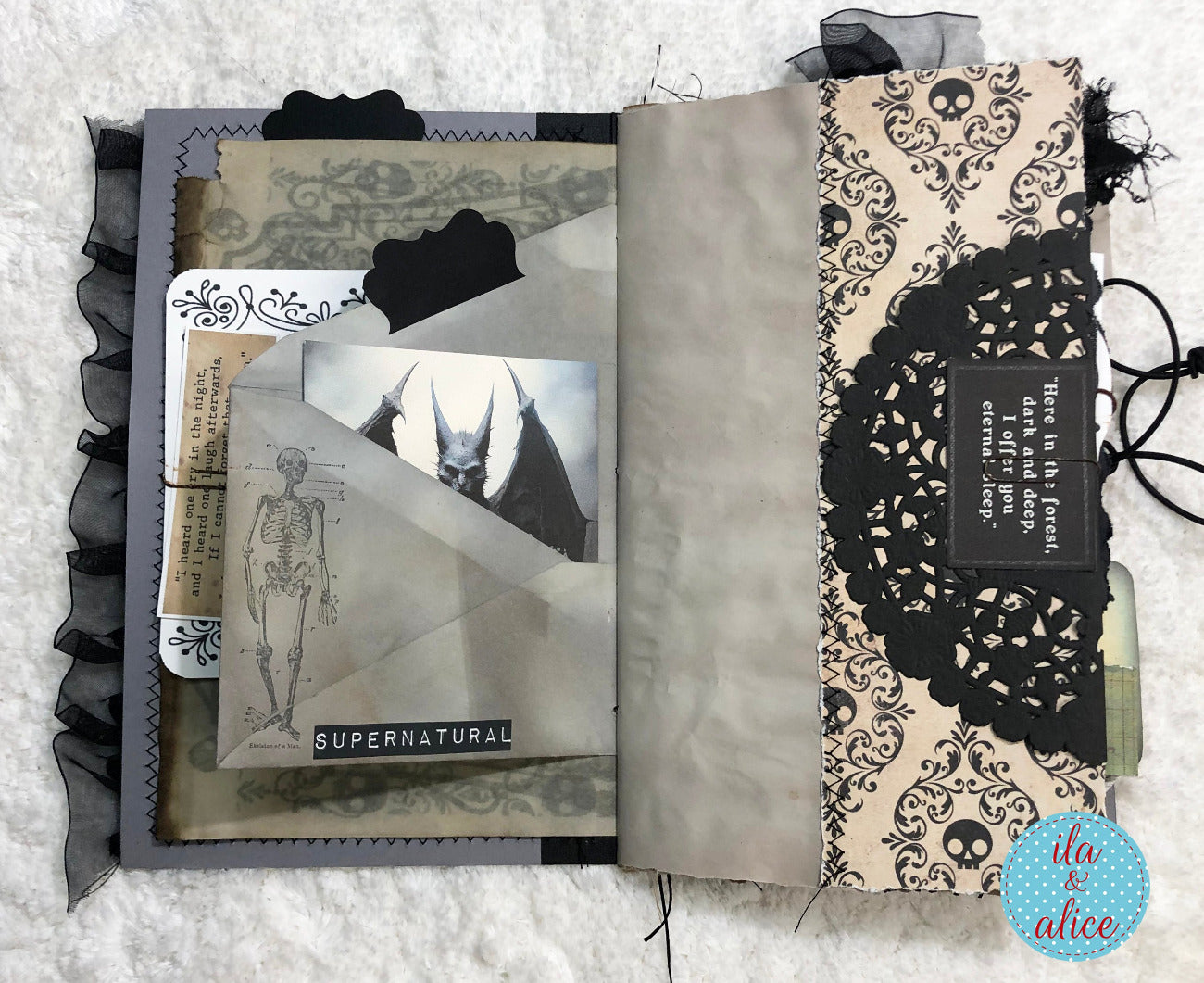 An open handmade scrapbook journal or art book displayed on a white textured fabric background. The left page features layered gothic elements including black ruffled trim, a skeleton illustration, a bat photo, and a 'SUPERNATURAL' label.