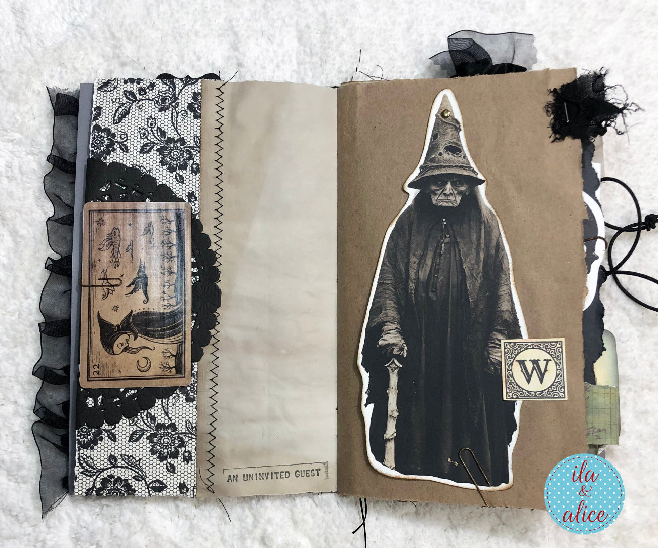 A handmade gothic scrapbook journal or art book shown open on a white textured fabric background. The pages feature black lace, brown kraft paper, black ruffled trim, a vintage tarot card, a black-and-white illustration of a hooded figure, and a small decorative seal.
